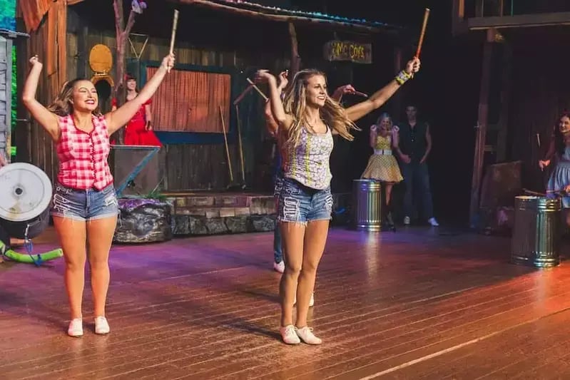 Hatfield & McCoy Dinner Feud performers dance on a wooden stage holding sticks with barrels, wearing costumes and boots.