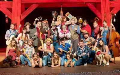 Cast of the Hatfield & McCoy Dinner Feud show posing on a rustic stage with red beams and wearing colorful costumes