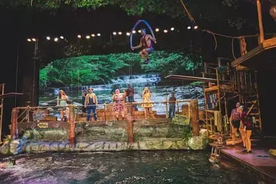 Performer on platform dives into swimming hole on stage while other cast members watch, with rustic set and greenery