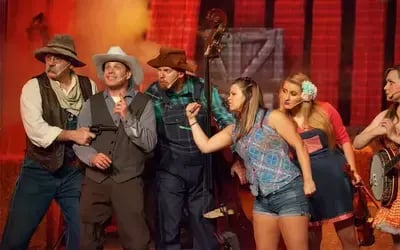 Performers in Western outfits dancing at Hatfield & McCoy Dinner Feud while musicians play banjo and guitar on rustic stage