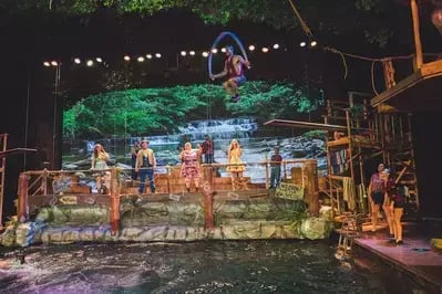Performers on the Hatfield & McCoy Dinner Feud stage around a water tank with rustic set pieces, greenery and lights.