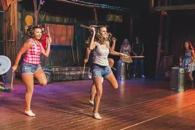 Two dancers performing on stage at Hatfield & McCoy Dinner Feud amid colorful lights and musicians in the background.